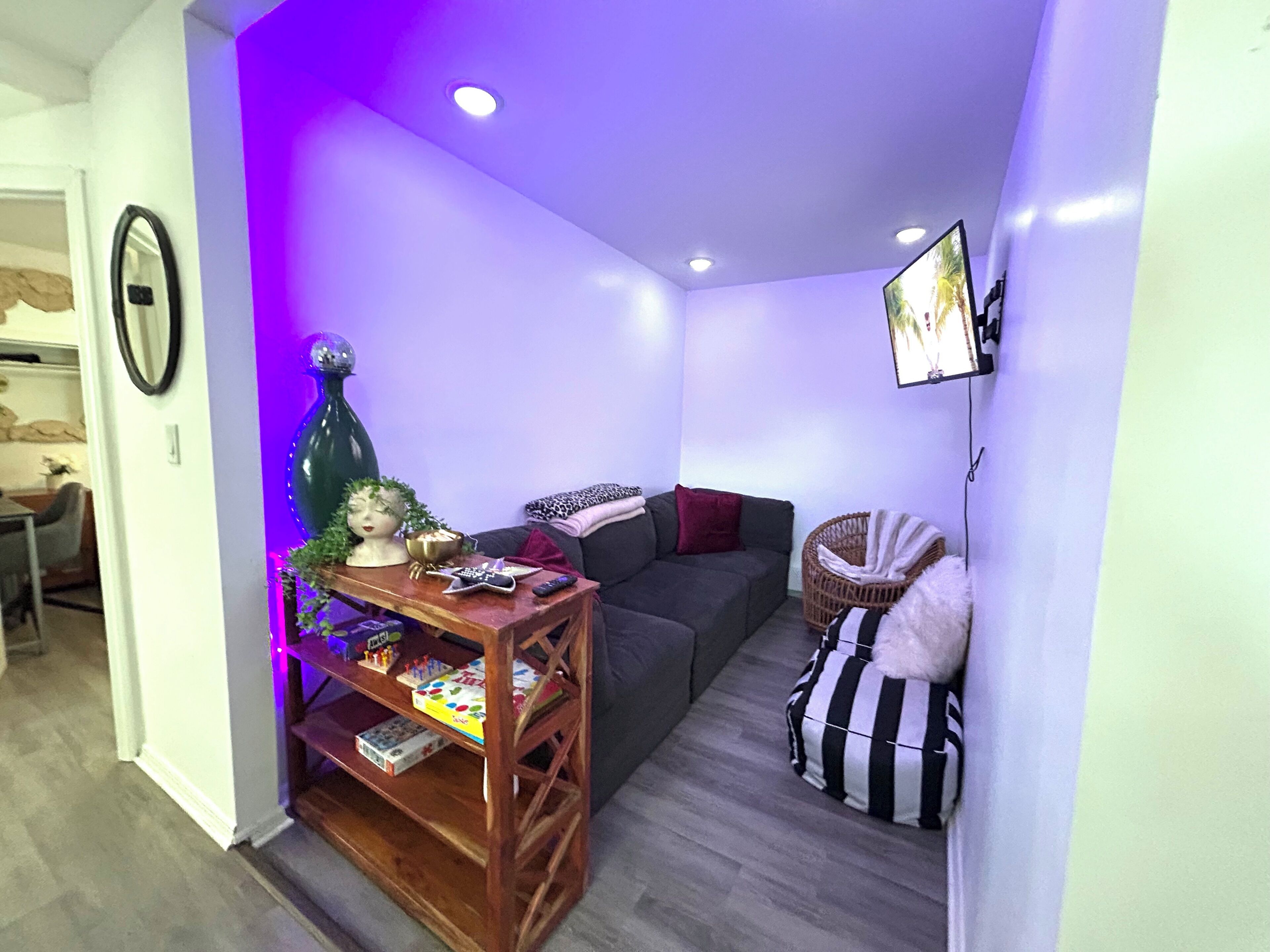 A compact living space with a sofa, a small table with items, a wicker chair, and a wall-mounted TV.