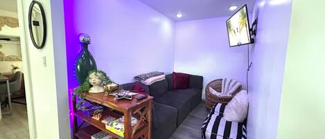 A compact living space with a sofa, a small table with items, a wicker chair, and a wall-mounted TV.
