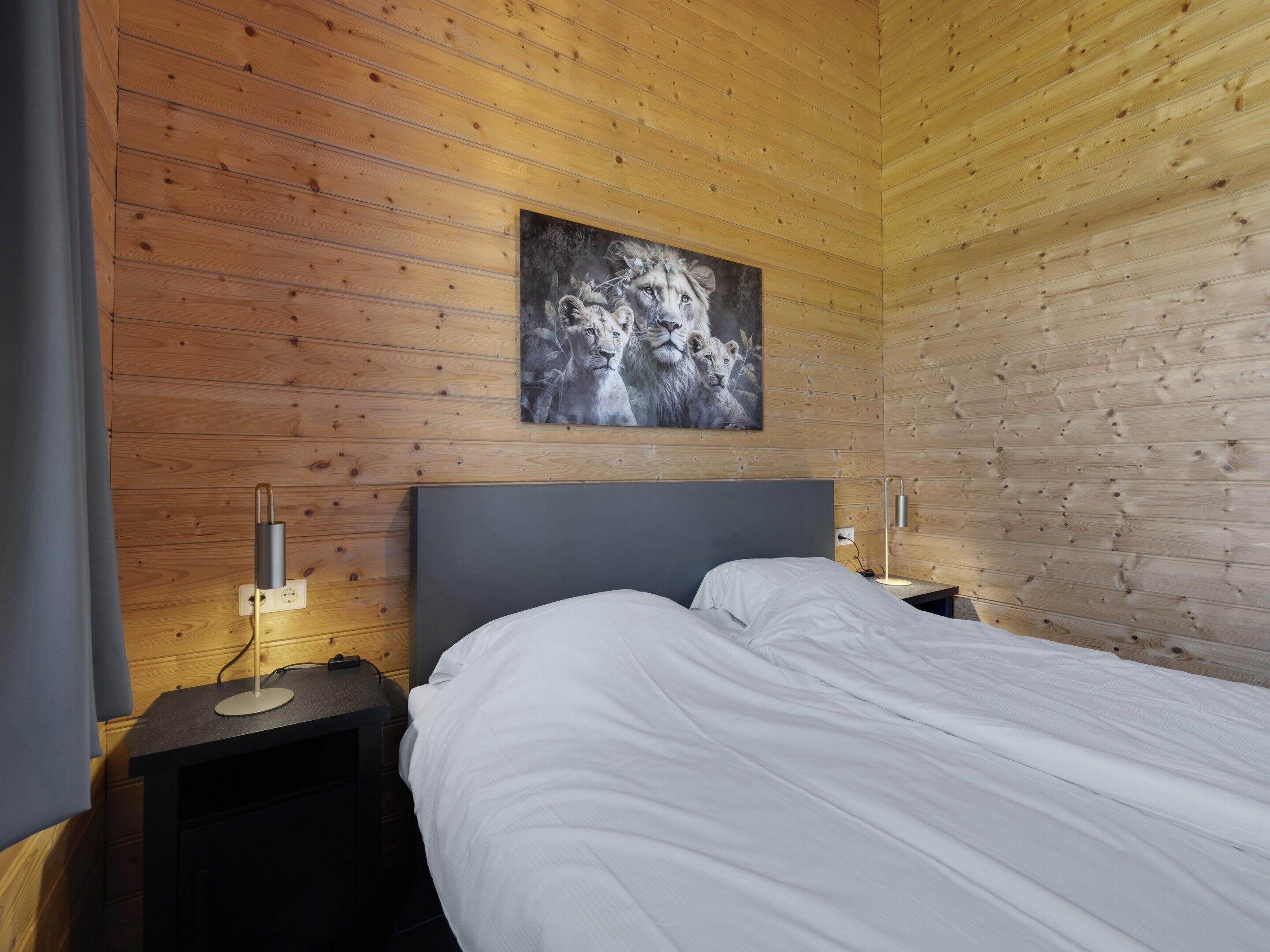 Ein Zimmer mit Holzwänden, einem Bett, einem Nachttisch, einer Lampe und einem Gemälde von Löwen.