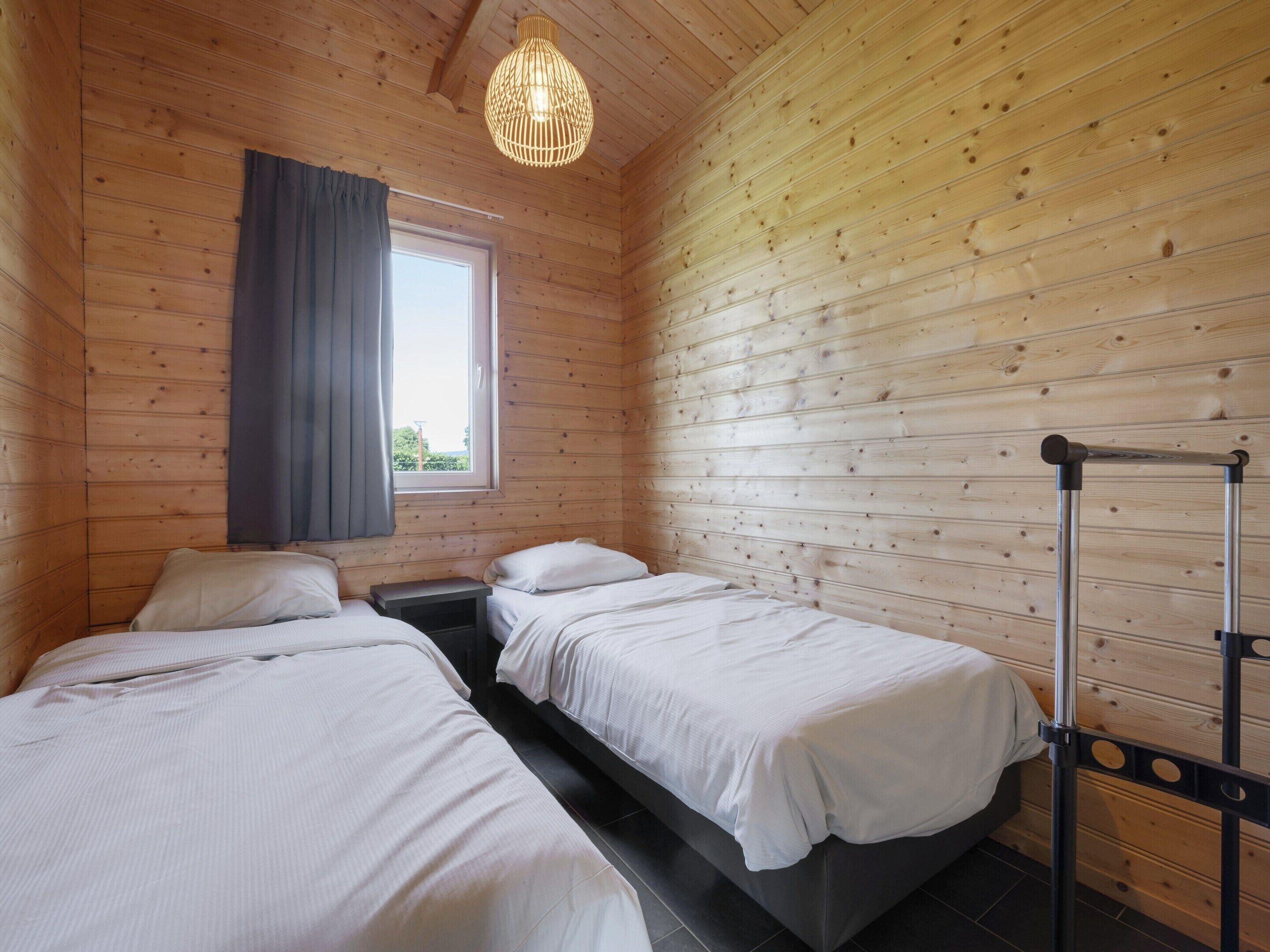 Ein Zimmer einer Holzjagdhütte mit zwei Betten, einem Fenster mit Vorhängen, einer Hügelampe und einem Metallständer.
