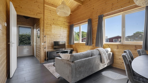 Interior de una cabaña de madera con sofá, televisor, mesa de comedor y ventanas amplias.