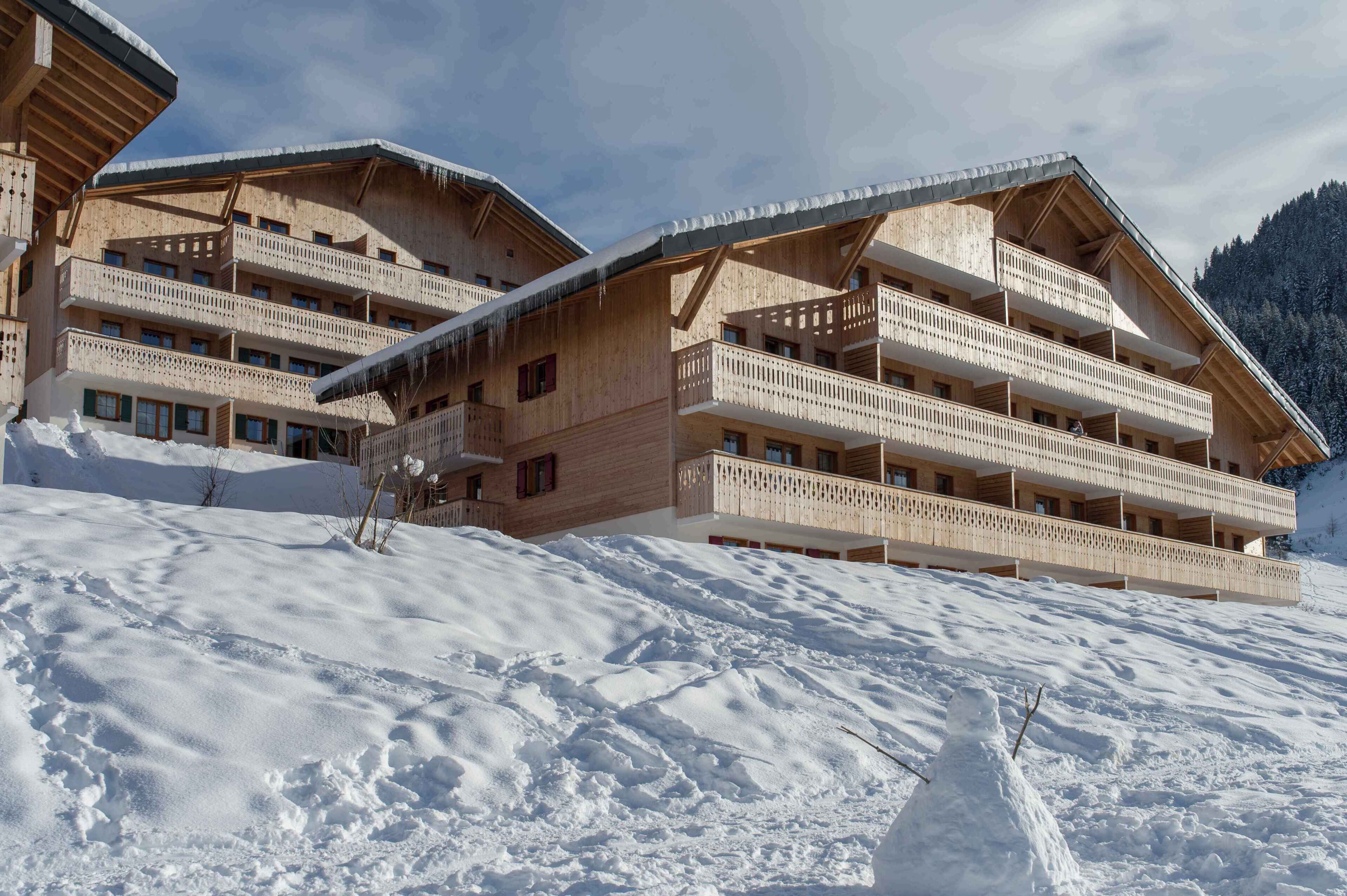 Ein Chalet-Hotel aus Holz mit mehreren Balkonen vor einer verschneiten Bergkulisse.