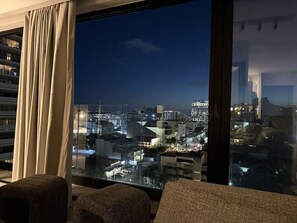 Ein Zimmer mit großem Fenster und Blick auf eine nächtliche Stadtlandschaft.