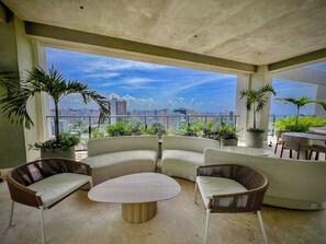 Un espace de détente extérieur moderne, doté d’un canapé et de fauteuils aux lignes courbes, agrémenté de plantes en pot et offrant une vue sur le paysage urbain.