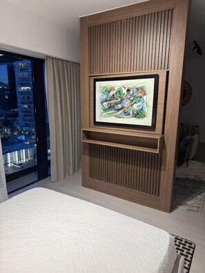 Ein Schlafzimmer mit einem Bett, einem an der Wand befestigten Fernseher und Blick auf die Stadt durch ein Fenster.