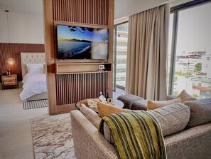 A modern hotel room with a large flat-screen TV mounted on a wooden panel, a comfortable sofa set, a bed with white linens, and a city view through large windows. - MStudio 1Bed in heart of Piantini, Santo Domingo (Santo Domingo)