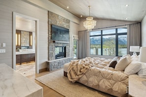 A bedroom with a stone fireplace, a large bed, a TV mounted on the wall, and a view of mountains through large windows.
