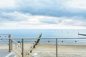 A beach view with a wooden pier extending into calm waters, a metal railing, and a sandy beach area.