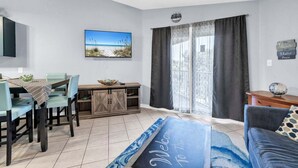 A living room with a dining area, a TV mounted on the wall, and a view of the beach through the window. - Private Beach Access, Sleeps 6, Pool & More! (Miramar Beach)