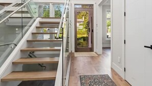 Et moderne interiør med en glas trappe, trægulv og en dør, der fører til et udendørs område med grønne planter.