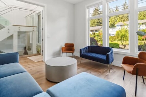 Ein modernes Wohnzimmer mit einem blauen Sofa, einem runden Couchtisch und Blick auf einen Garten durch große Fenster.