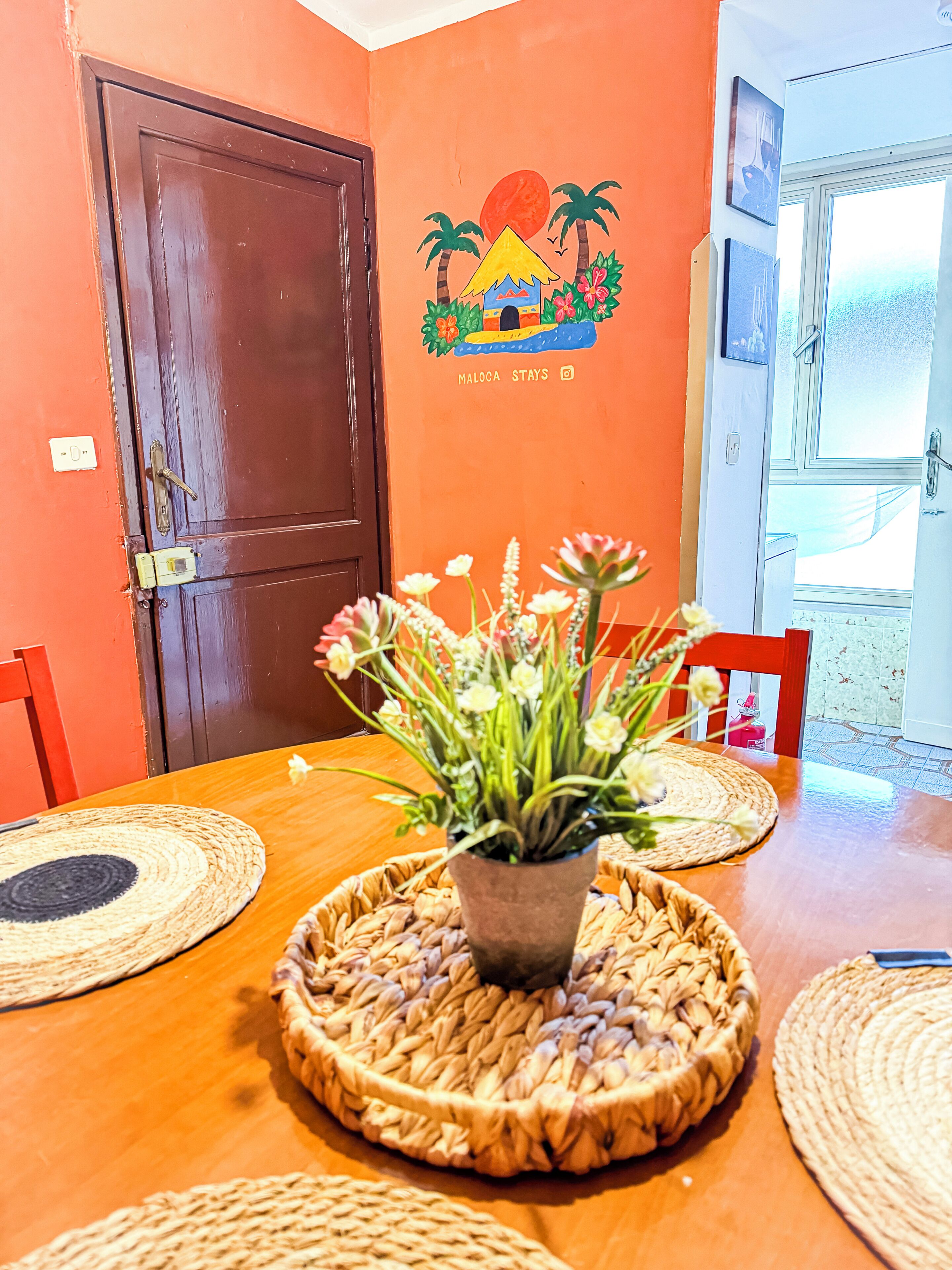 Un coin repas avec une table en bois, des sets de table en fibres tressées et une plante en pot.