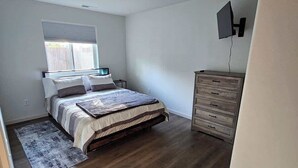 A bedroom with a bed, a dresser, a TV mounted on the wall, and a window with blinds.