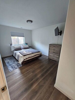 A bedroom with a bed, a dresser, a TV mounted on the wall, and a window with blinds.