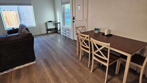 Ein geräumiges Zimmer mit Holzboden, einem Esstisch mit Stühlen, einer Couch und einem Fenster mit Jalousien.
