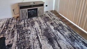 Ein Zimmer mit montiertem Fernseher, einer Holztruhe zur Unterhaltungselektronik und einem gemusterten Teppich.
