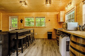 Interior de una cabaña de madera con área de cocina, asientos de bar y un ventanal grande.