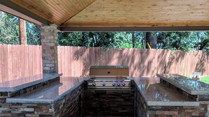 Une cuisine extérieure équipée d’un barbecue, de plans de travail en pierre et d’un ventilateur de plafond.