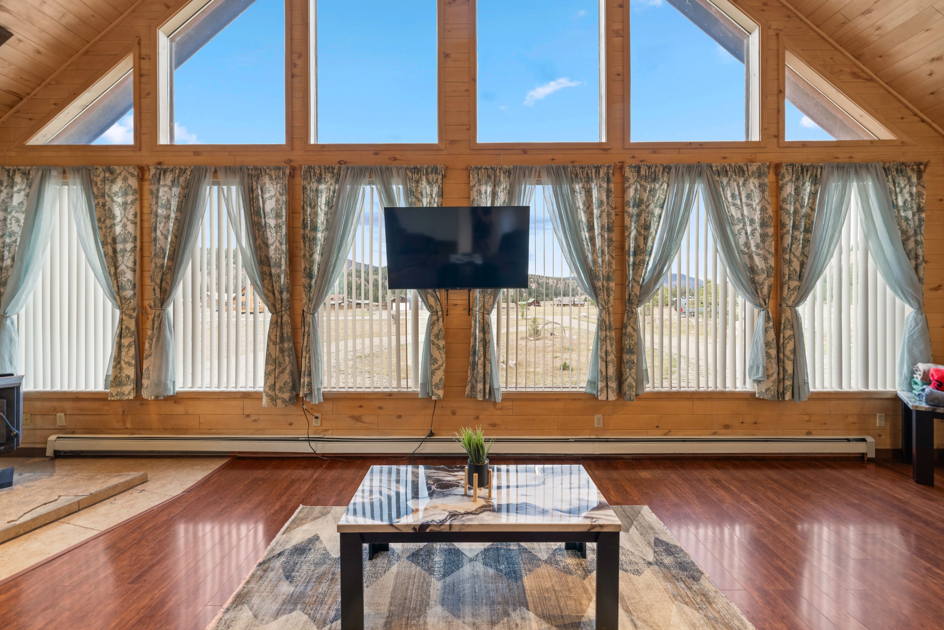 Ein Zimmer mit einem großen Fernseher, Holzböden, einem Tisch mit einer Vase und großen Fenstern mit Vorhängen.
