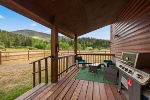 Une terrasse en bois avec un barbecue et du mobilier d’extérieur, donnant sur une zone verdoyante avec des arbres et des collines.