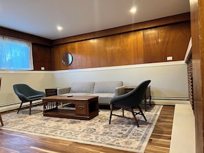 Una sala de estar con un sofá, dos sillones, una mesa de centro, un tapete, paneles de madera, un espejo y una escalera.