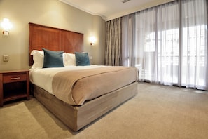 A neatly made bed with a wooden headboard, a nightstand, and a large window with sheer curtains. - The Raphael Penthouse Suites (Johannesburg)