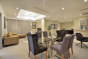 A modern living room with a glass-top dining table, plush seating, and a large window with curtains. - The Raphael Penthouse Suites (Johannesburg)