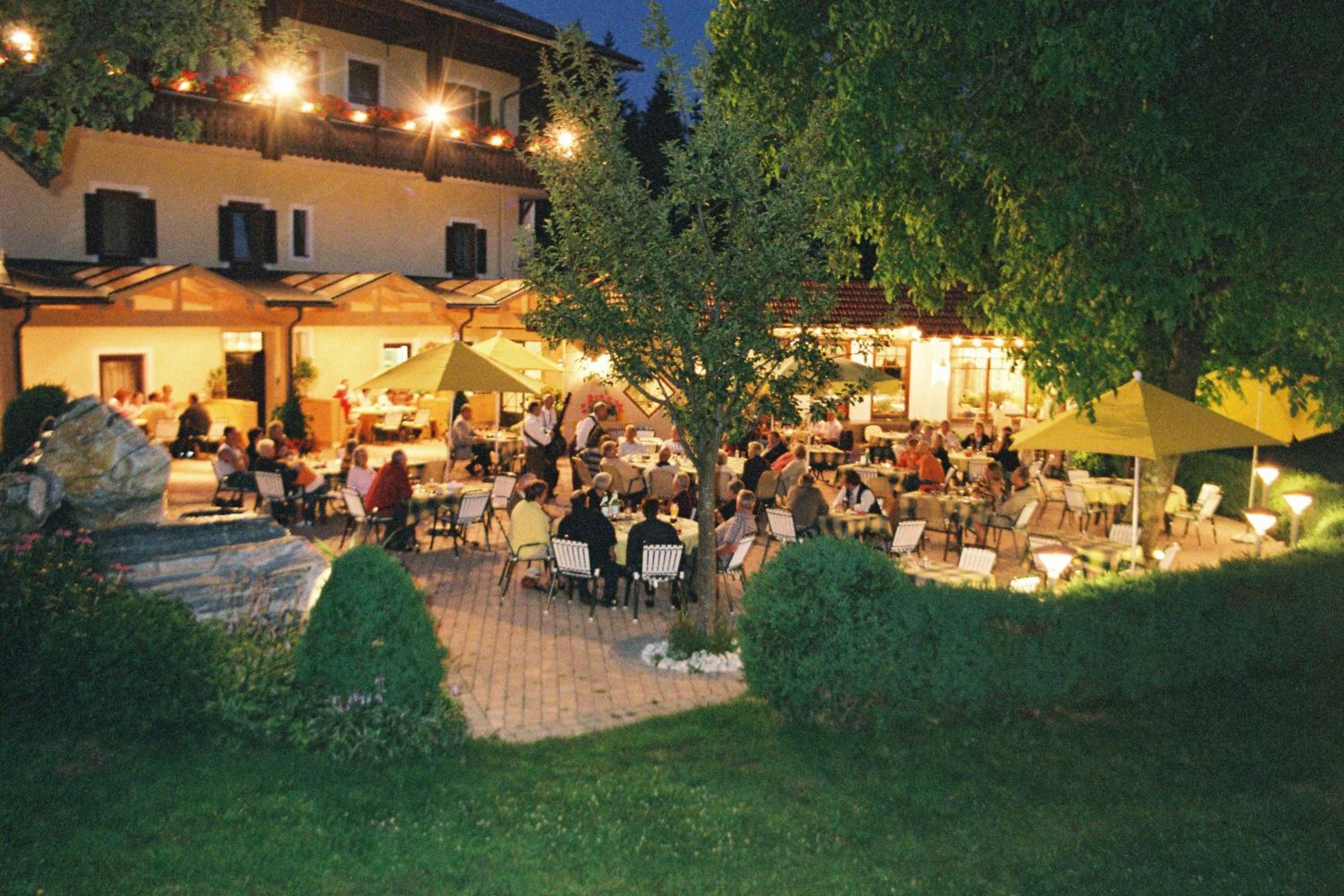 An outdoor dining area with tables and chairs, surrounded by greenery and a building with multiple windows.