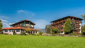 Een hotel in traditionele Alpenstijl, met meerdere balkons met bloembakken, voor een achtergrond van groene velden en bergen.
