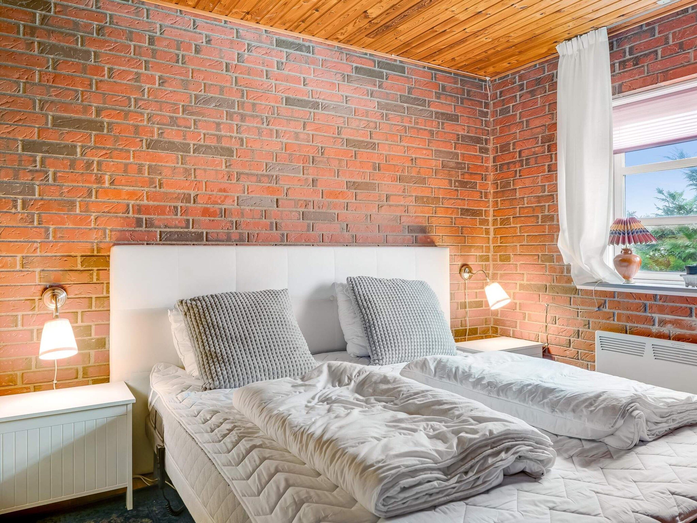A bedroom with a brick wall, a white headboard, two bedside lamps, a window with a curtain, and a bed with white bedding.