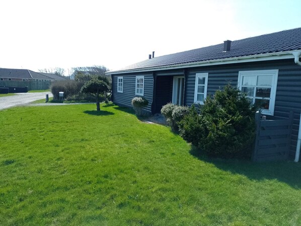 Ett envåningshus med en mörk fasad, en ytterdörr och en liten trädgård framför huset med ett träd och några buskar.