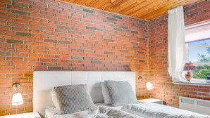 Ein Schlafzimmer mit Backsteinwand, weißem Kopfteil, zwei Nachttischlampen, einem Fenster mit Vorhang und einer Heizung.