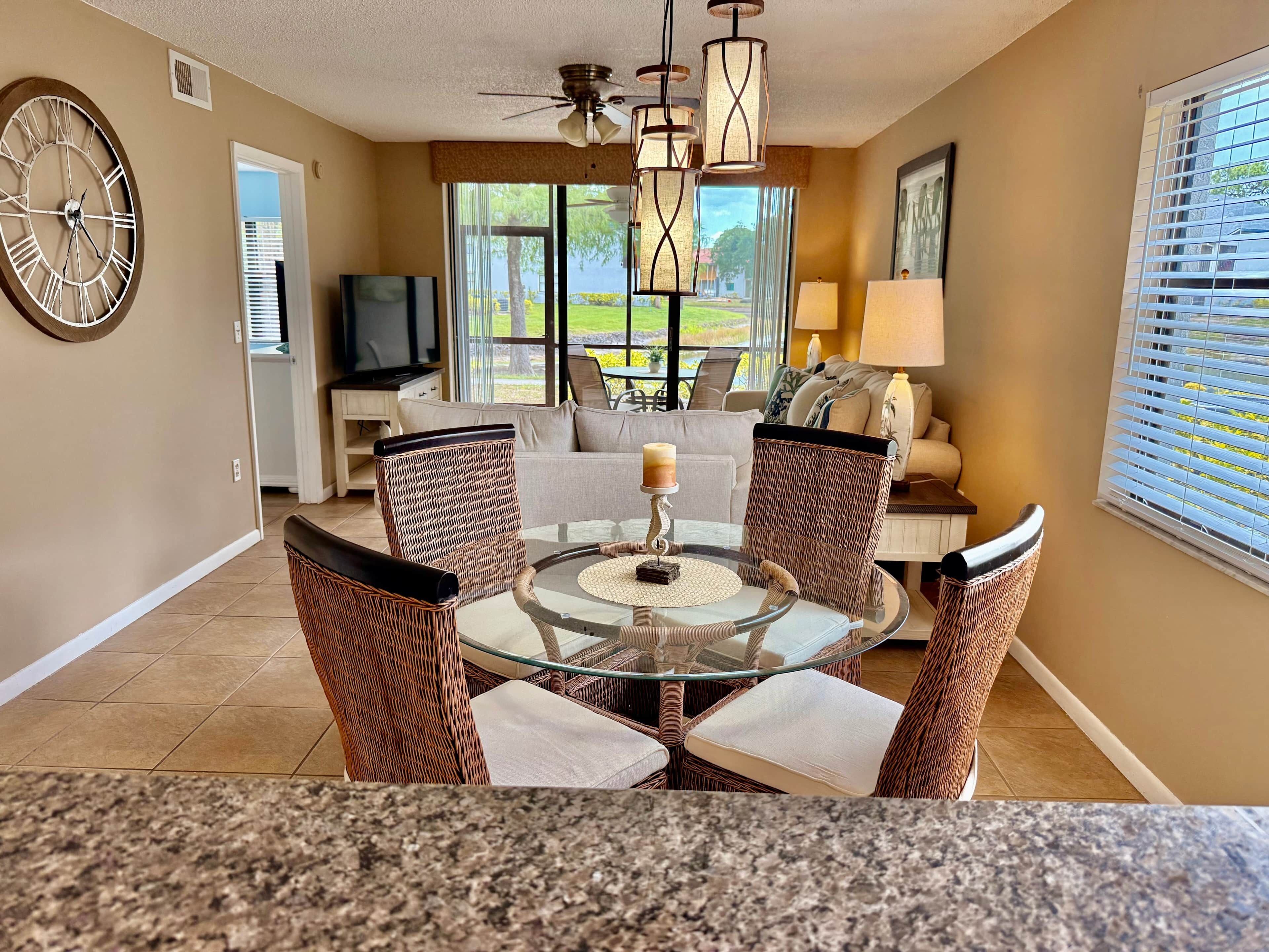 A dining area with wicker chairs, a glass table, a clock, and a TV in the background.