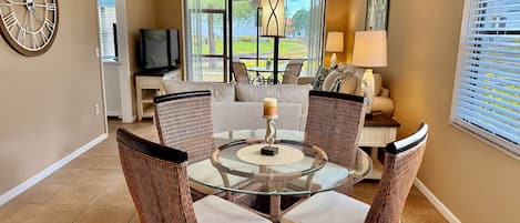 A dining area with wicker chairs, a glass table, a clock, and a TV in the background.