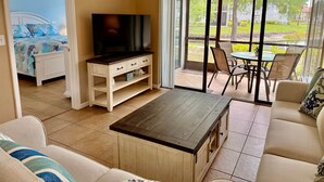 Un salon équipé d’un téléviseur, d’un canapé, d’une table basse et offrant une vue sur un jardin grâce à de grandes fenêtres.