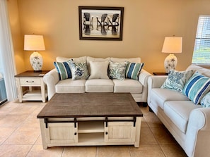 Ein Wohnzimmer mit einem beigefarbenen Sofa, zwei Beistelltischen mit Lampen, einem Couchtisch und einem gerahmten Bild an der Wand.