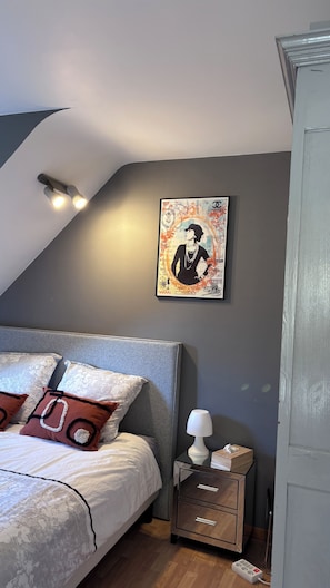 Ein Schlafzimmer mit Bett, Kissen, einem Nachttisch, einer Lampe, einem Bilderrahmen und Deckenleuchte.