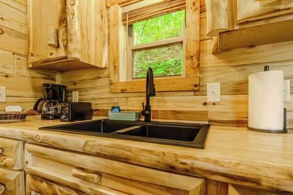 A wooden cabin kitchen with a double sink, wooden cabinets, a window with a view of greenery, and a basket with a bottle.