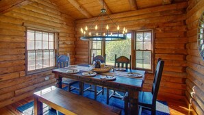 A wooden cabin interior with a dining table set for four, large windows, and a hanging light fixture.