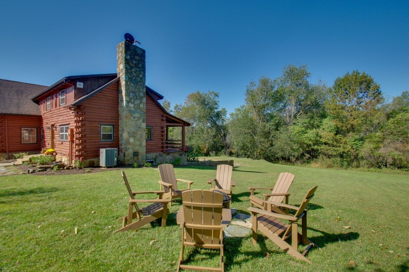 A log cabin with a stone chimney, surrounded by a grassy yard with Adirondack chairs.