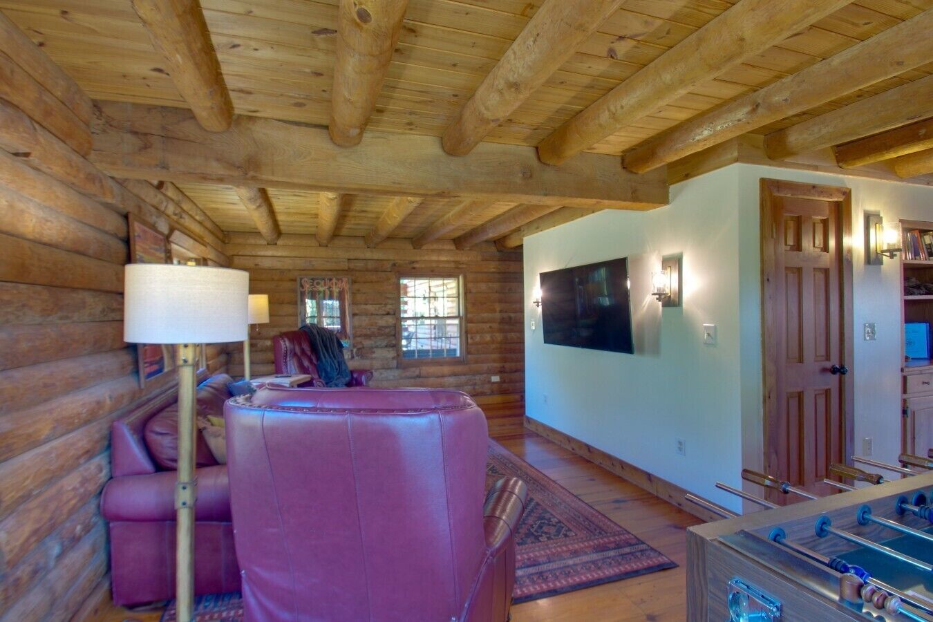 A log cabin interior with a sofa, a television, a foosball table, and a door leading to another room.