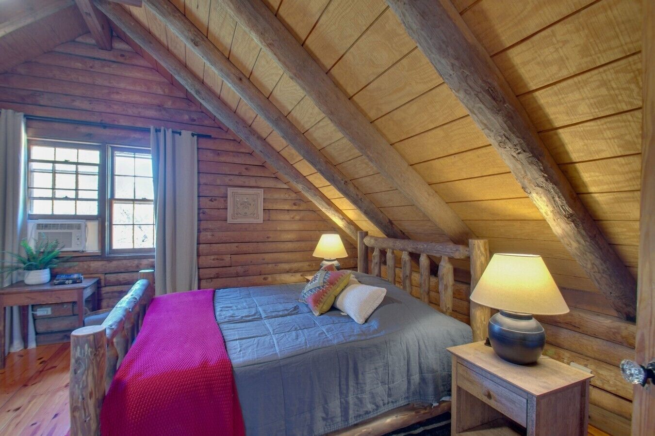 A cozy attic bedroom with a wooden ceiling, a bed with pillows, a nightstand with a lamp, and a window with curtains.