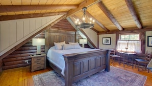 A log cabin bedroom with a large bed, wooden beams, a chandelier, a desk, and a window.