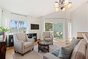 Ein modernes Wohnzimmer mit einer Couch, Sesseln, einem Couchtisch und einem Fernseher. Ein Balkon bietet Blick auf Palmen.