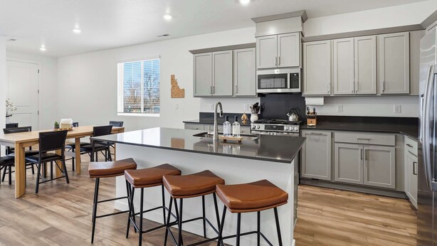 Een moderne keuken met grijze kastjes, een centraal eiland met barkrukken, een eethoek met een houten tafel en stoelen, en een groot raam met jaloezieën.