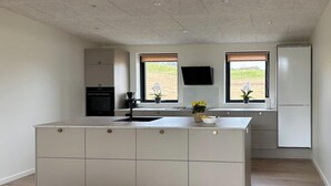 A modern kitchen with a central island, two windows, a built-in oven, and a flat-screen TV. - House on large nature plot with sea view and spa (Broager)