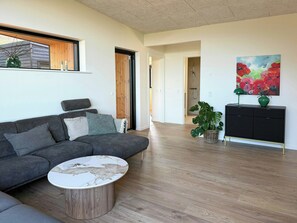 A modern living room with a grey sectional sofa, a round coffee table, a black console table with a green vase, and a large floral painting on the wall. - House on large nature plot with sea view and spa (Broager)