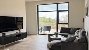 A modern living room with a large TV, a sofa, a coffee table, and a view through a large window. - House on large nature plot with sea view and spa (Broager)