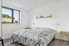 A bedroom with a large bed, a window with a view of the outdoors, a wooden shelf, and a desk with a chair. - House on large nature plot with sea view and spa (Broager)