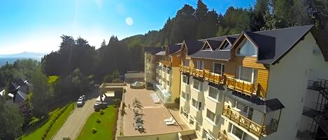 A multi-story hotel with a balcony on the upper floors, surrounded by greenery and a view of the landscape.